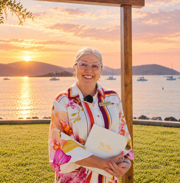Leanne at sunset beneath a wooden arbour overlooking the Whitsundays, holding a Whitsunday Love Stories ceremony folder