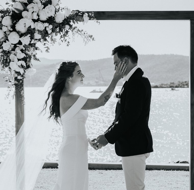 Bride and groom exchanging vows under a floral arbour beside the water in Airlie Beach