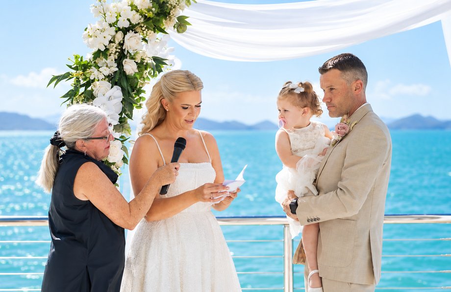 Bride reading her vows with Leanne holding the microphone, groom holding their daughter, overlooking the Coral Sea